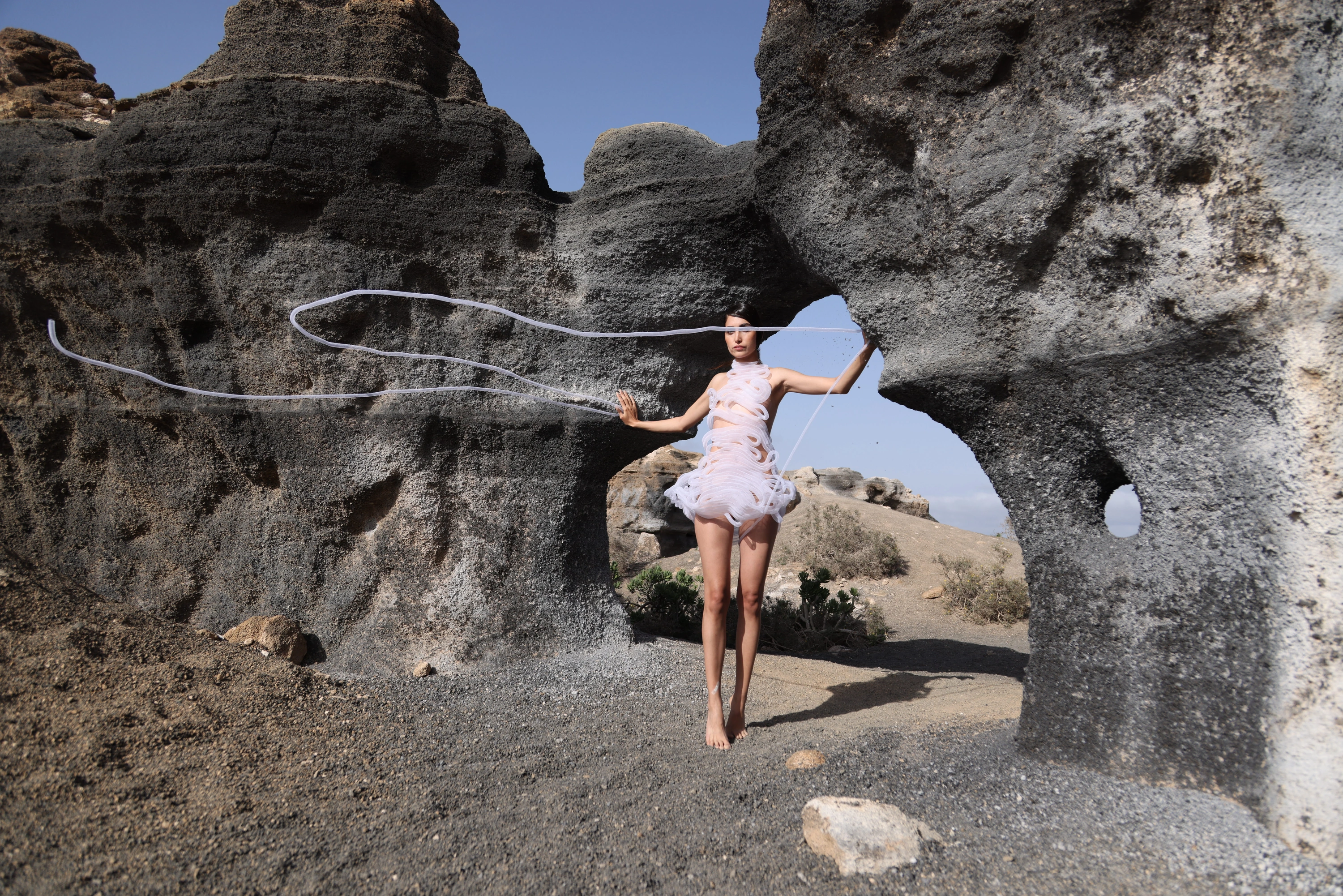 Sculptural pale textile garment with trailing tubes framed by volcanic rock